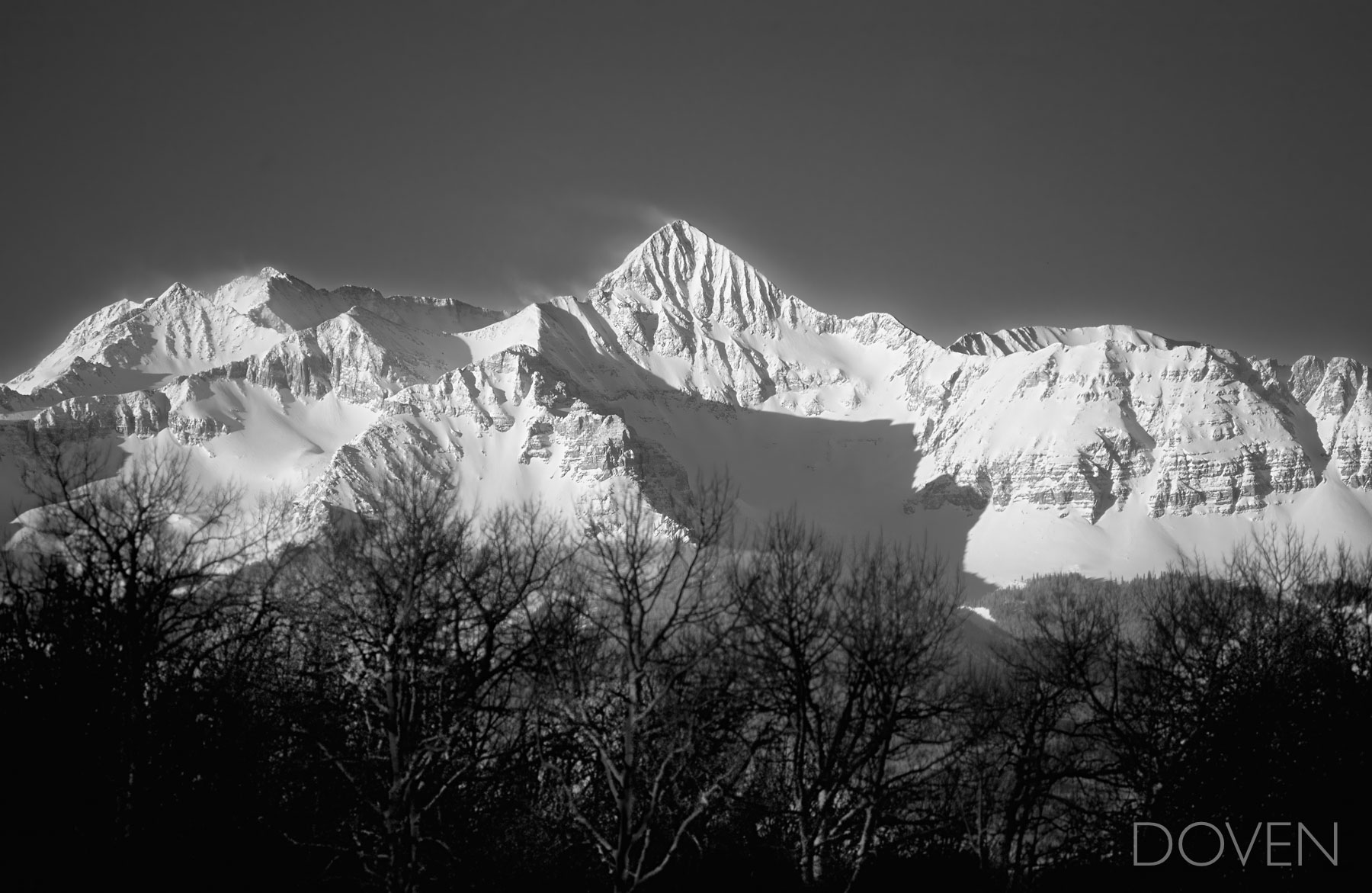 Black trees and gray sky frame white majestic mountains as dawn breaks on the steep and snow covered slopes of the iconic Mt...
